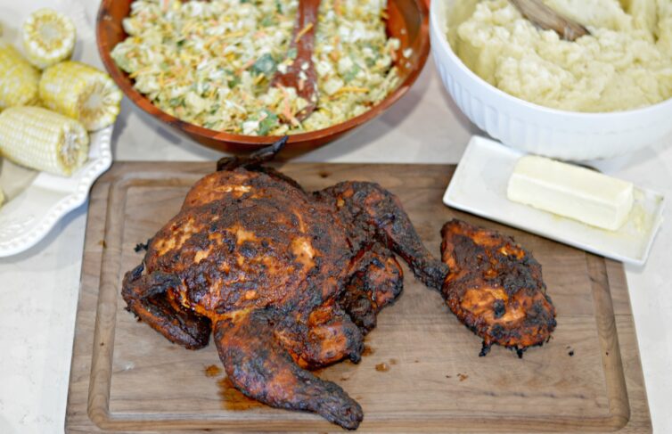 Smoked whole chicken on a cutting board with salad, corn and mashed potatoes in the background