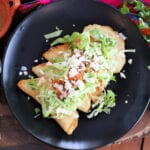 Mexican empanadas on a black plate and topped with pico de gallo, lettuce, and crema