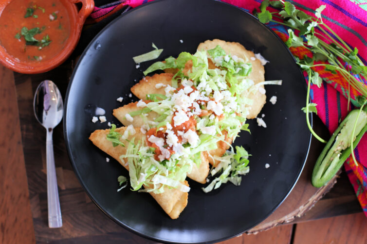 Mexican empanadas on a black plate and topped with pico de gallo, lettuce, and crema